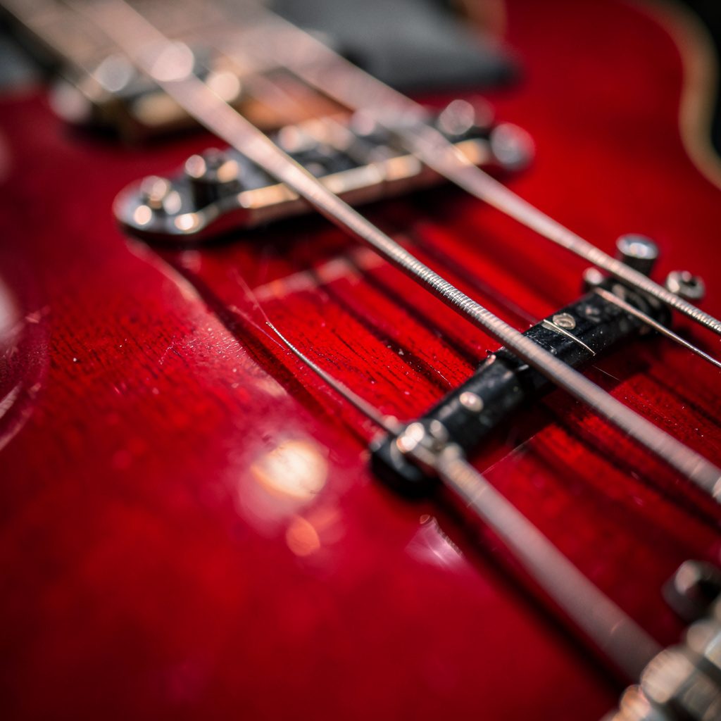 Close-up photograph of two bass guitars leaning against each other, with warm wood browns and deep cherry reds set against a moody charcoal studio backdrop.